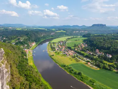 Scenic panoramic view from the Bastei cliffs over the Elbe River valley with sandstone rocks, lush forests, rural village rooftops, distant mountain ranges under a vibrant summer sky. Saxony, Germany
